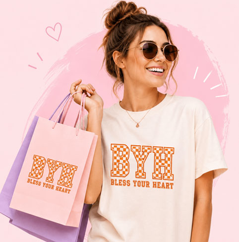 A woman wearing round sunglasses and a cream graphic t-shirt with orange 'BYU BLESS YOUR HEART' lettering, holding pink and purple shopping bags against a pink graphic background.