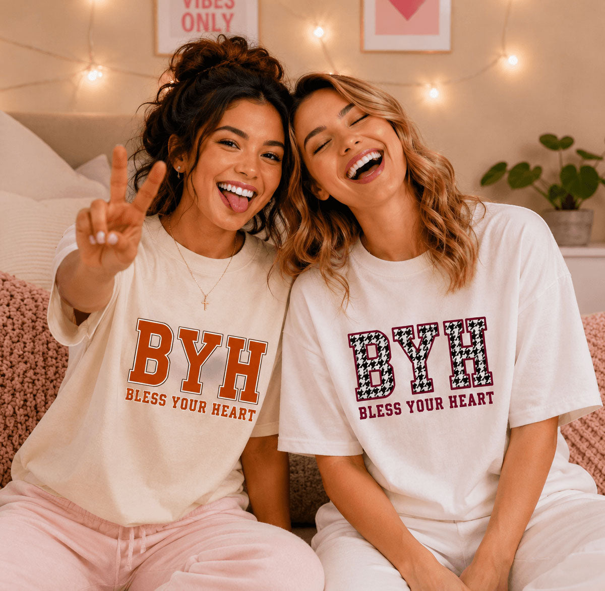 Two women sitting together on a couch wearing matching white graphic t-shirts with 'BYH BLESS YOUR HEART' printed on the front in orange and black-and-white patterned lettering, in a cozy bedroom setting with warm string lights.