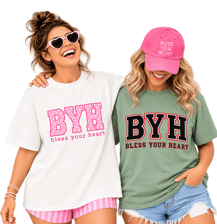 Two women posing together wearing graphic t-shirts with 'BYH' and 'Bless Your Heart' text. The woman on the left wears a white t-shirt with pink text and pink striped shorts, while the woman on the right wears a sage green t-shirt with black and red text and a pink cap.
